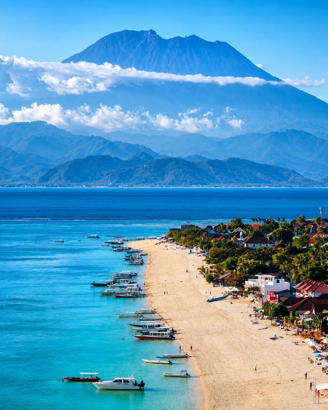 Boats lined up on a golden beach with a volcano rising across the turquoise water, Bali