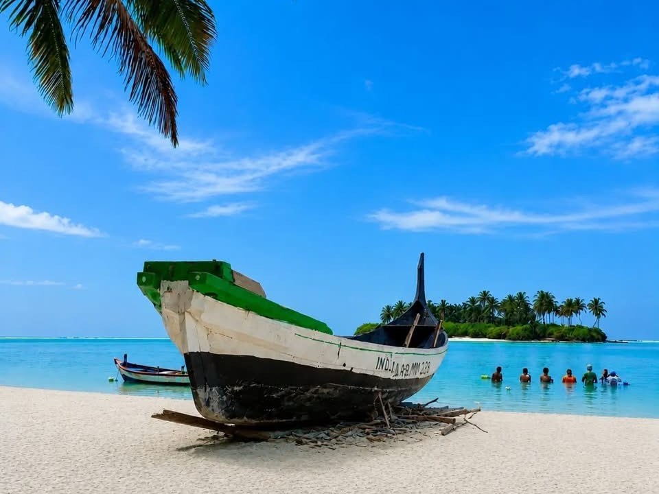 Traditional wooden fishing boat on white sand under a coconut palm, with an islet across the turquoise lagoon, Lakshadweep
