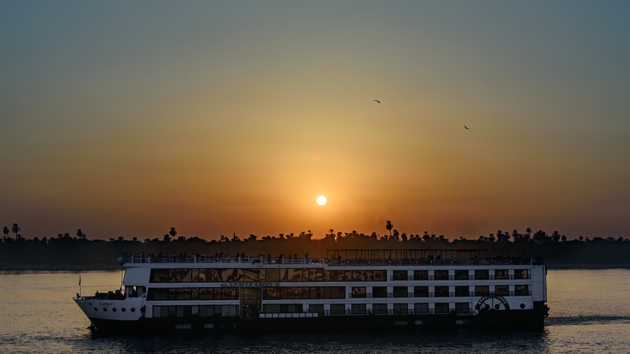 A Nile cruise boat sailing past a line of palm trees at sunset between Aswan and Luxor, Egypt