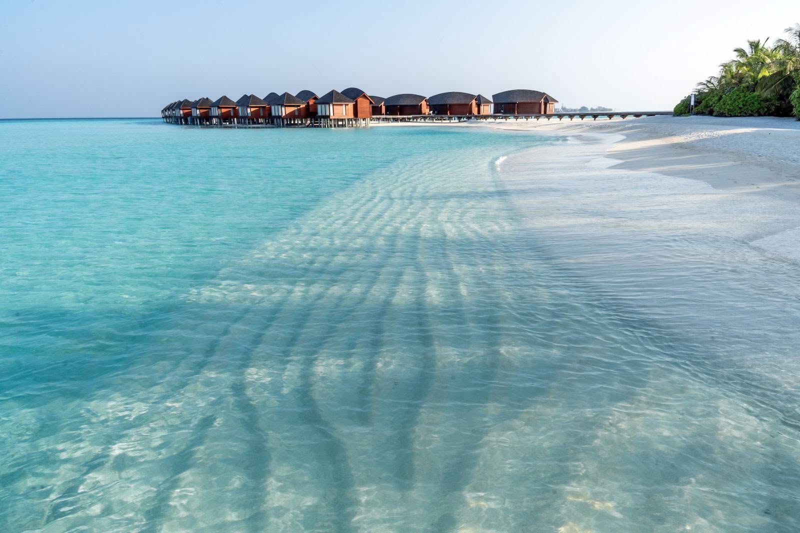 A row of overwater villas on stilts beside a pale turquoise lagoon and a white sand beach, Maldives