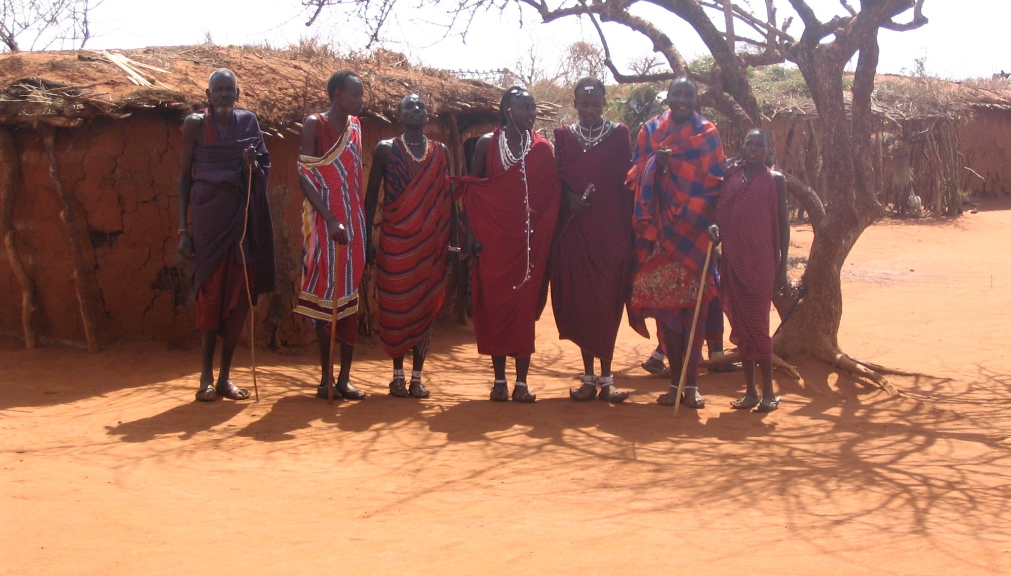 Maasai people in red and blue shukas standing in front of a mud-and-thatch hut in their village