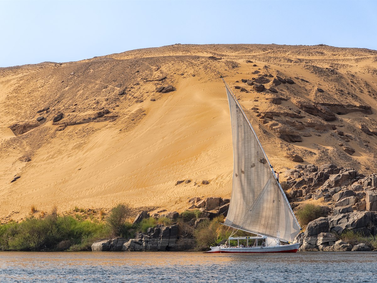 A white-sailed felucca on the Nile below a golden sand dune and dark granite rocks near Aswan, Egypt