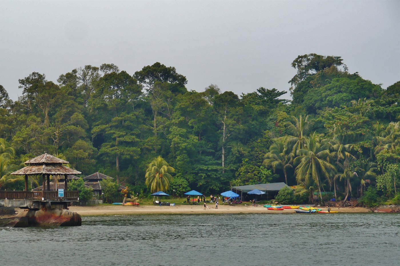 Colourful kayaks and blue beach umbrellas on Pulau Ubin's shore, palm trees and a wooden pavilion behind