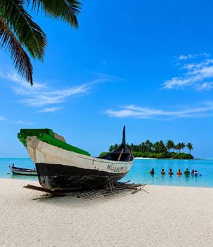 Traditional wooden fishing boat on white sand under a coconut palm, with an islet across the turquoise lagoon, Lakshadweep